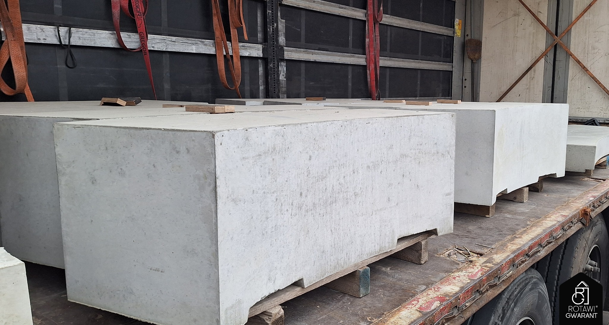 Szare, betonowe bloki ułożone na drewnianych paletach, zabezpieczone pasami transportowymi wewnątrz naczepy. Widoczne opony ciężarówki i logo producenta.