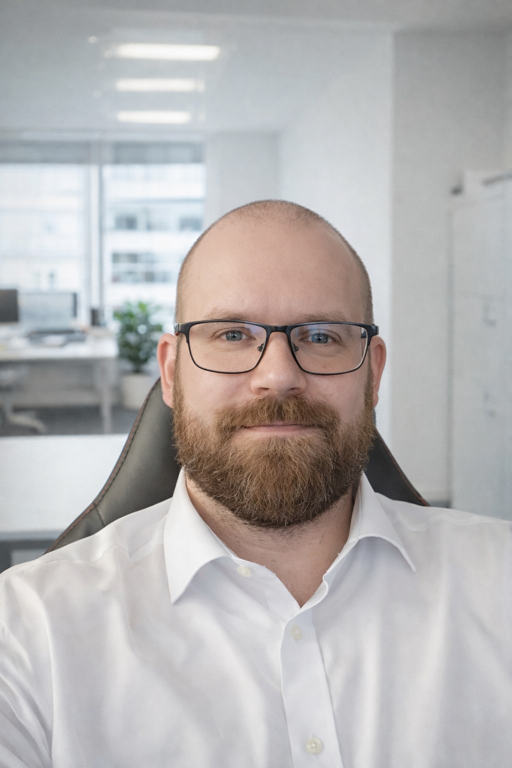 Portret mężczyzny z brodą w okularach, ubrany w białą koszulę, siedzącego na czarnym krześle biurowym. W tle jasne biuro z oknem i biurkami.