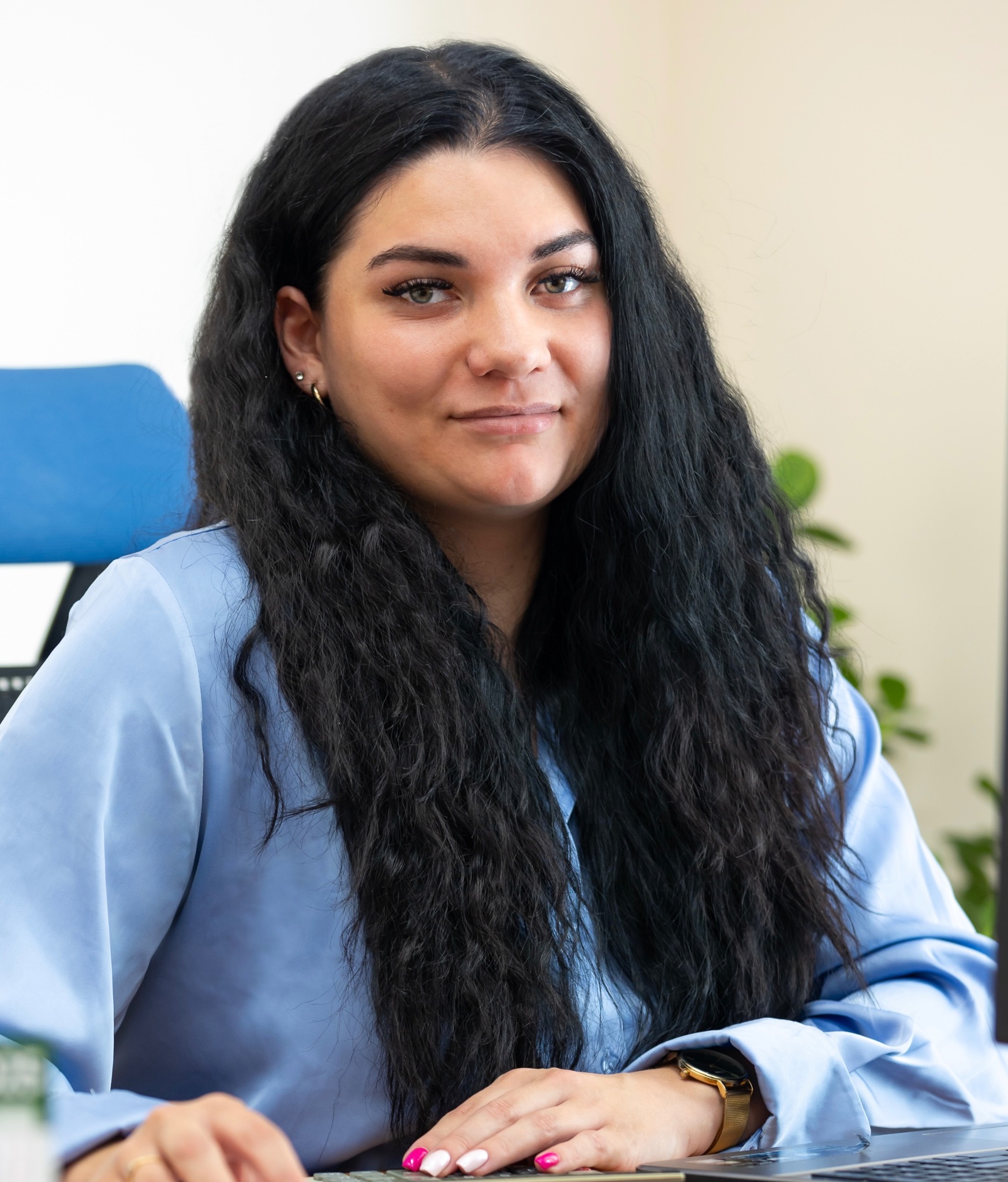 Portret kobiety z długimi, ciemnymi włosami w błękitnej koszuli, siedzącej przy biurku z laptopem i kalkulatorem. Uśmiechnięta, z zegarkiem na ręku.