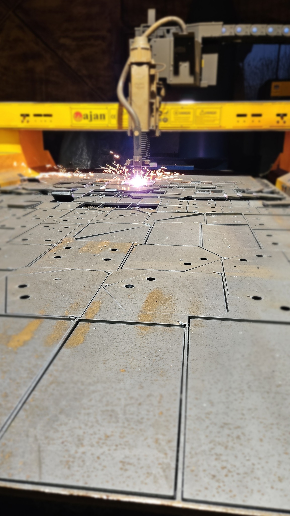 Automat tnący blachę na kwadratowe elementy z otworami. Iskry podczas cięcia. Perspektywa z dołu, widoczny fragment maszyny i pocięta blacha.