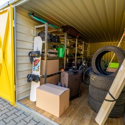 Własny Box - Wnętrze magazynu samoobsługowego: regały z torbami, walizkami, deską snowboardową, oponami i kartonami. Drewniana podłoga, żółte drzwi. Przechowywanie sezonowe.