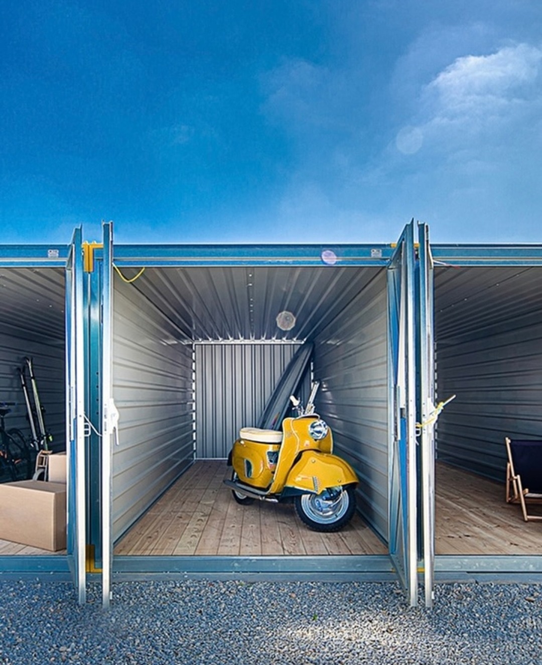 Otwarty kontener magazynowy z żółtym skuterem w środku, obok narty i kartony. Podłoga z desek, niebieskie niebo w tle. Dostępne boksy do wynajęcia.