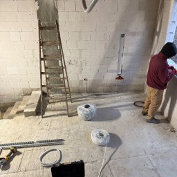 Electrius Jan Kazimierczak - Wnętrze w trakcie prac elektrycznych: instalacja przewodów w ścianie z bloków, widoczne kable w rolkach, drabina i narzędzia. Elektryk w trakcie pracy przy oknie.