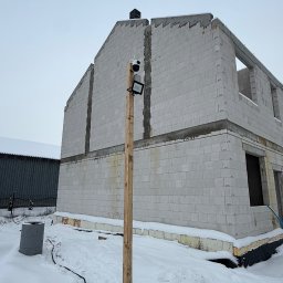 Electrius Jan Kazimierczak - Niedokończony budynek z betonu komórkowego w zimowej scenerii. Widoczny tymczasowy słup z zamontowaną kamerą i reflektorem. Kabel biegnie po śniegu.