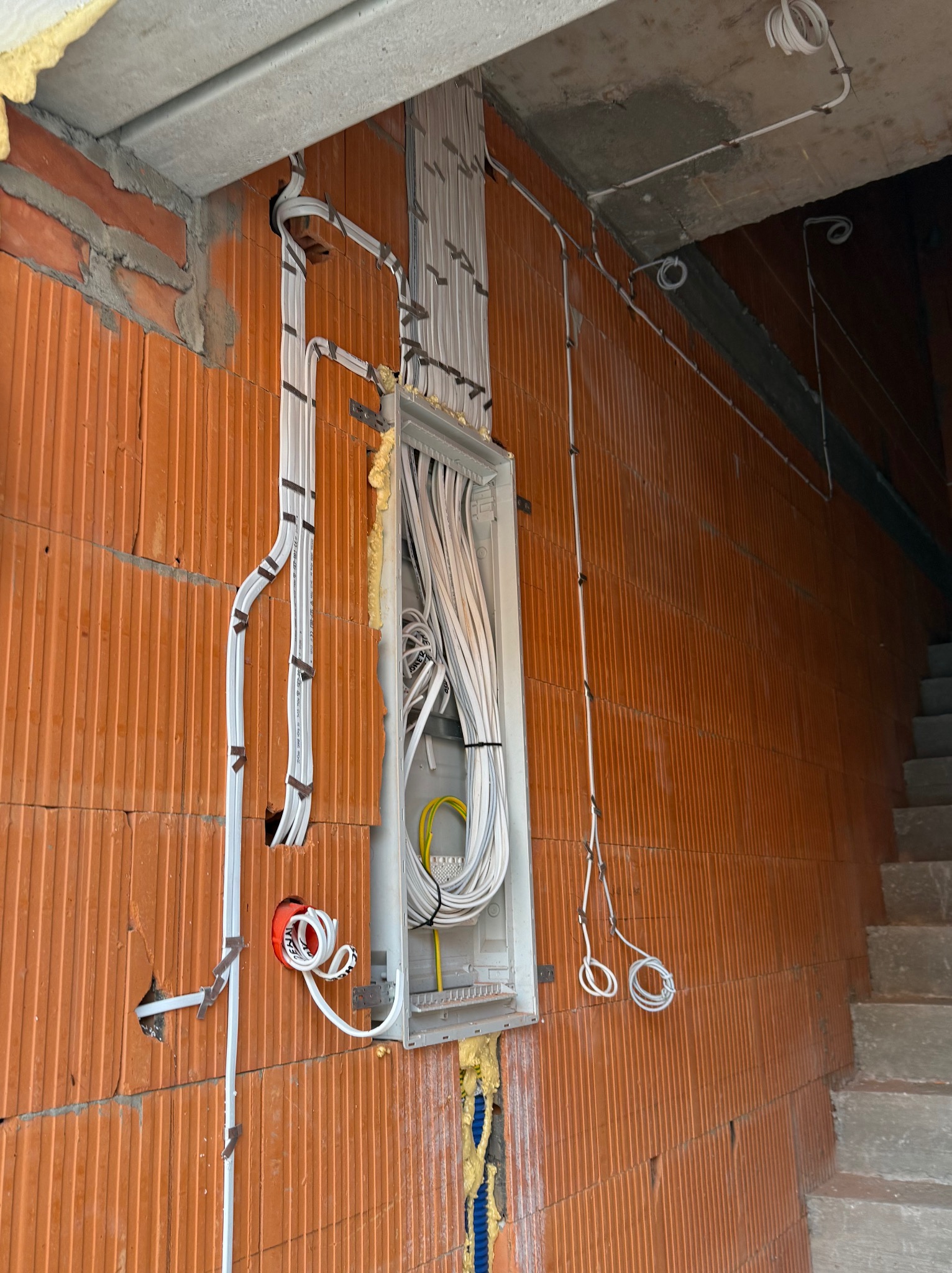 Rozprowadzona instalacja elektryczna w budynku w stanie surowym, widoczne kable w peszlach i puszka elektryczna na ceglanej ścianie.