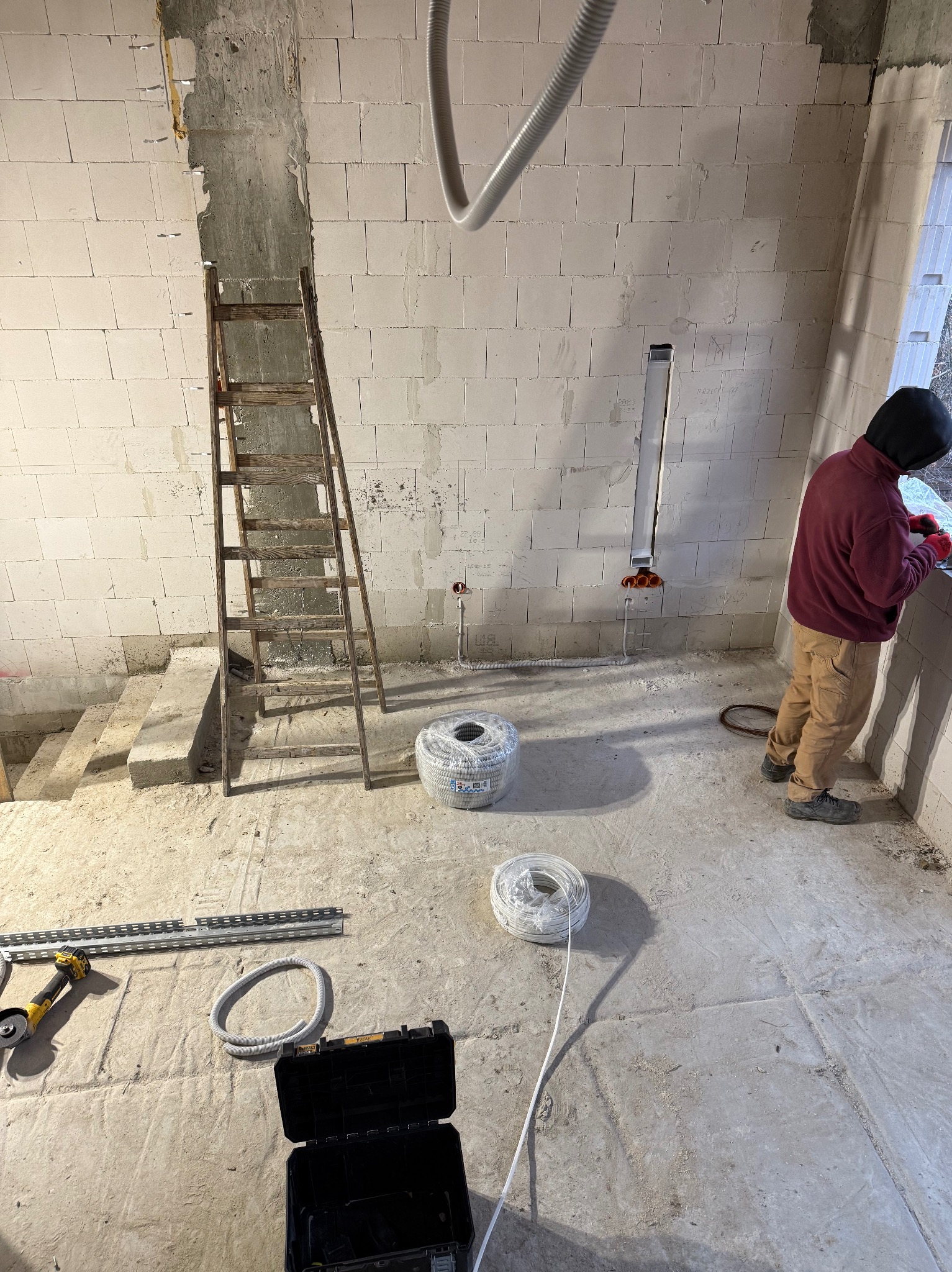 Wnętrze w trakcie prac elektrycznych: instalacja przewodów w ścianie z bloków, widoczne kable w rolkach, drabina i narzędzia. Elektryk w trakcie pracy przy oknie.