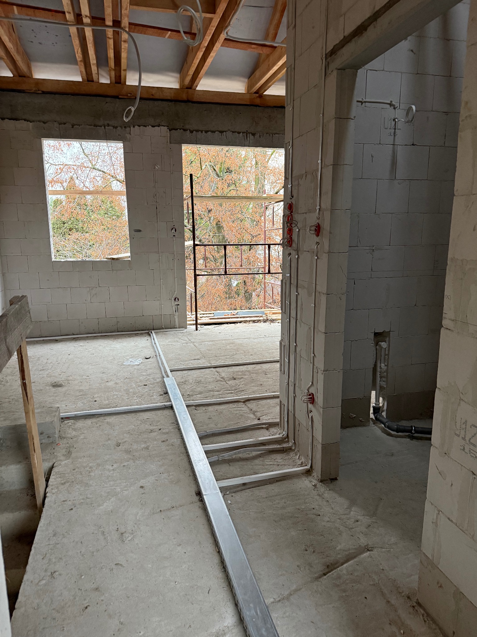 Wnętrze w stanie surowym z widoczną instalacją elektryczną w ścianach i podłodze. Kable prowadzone w peszlach, puszki elektryczne zamontowane. Widoczne okna i surowy strop.