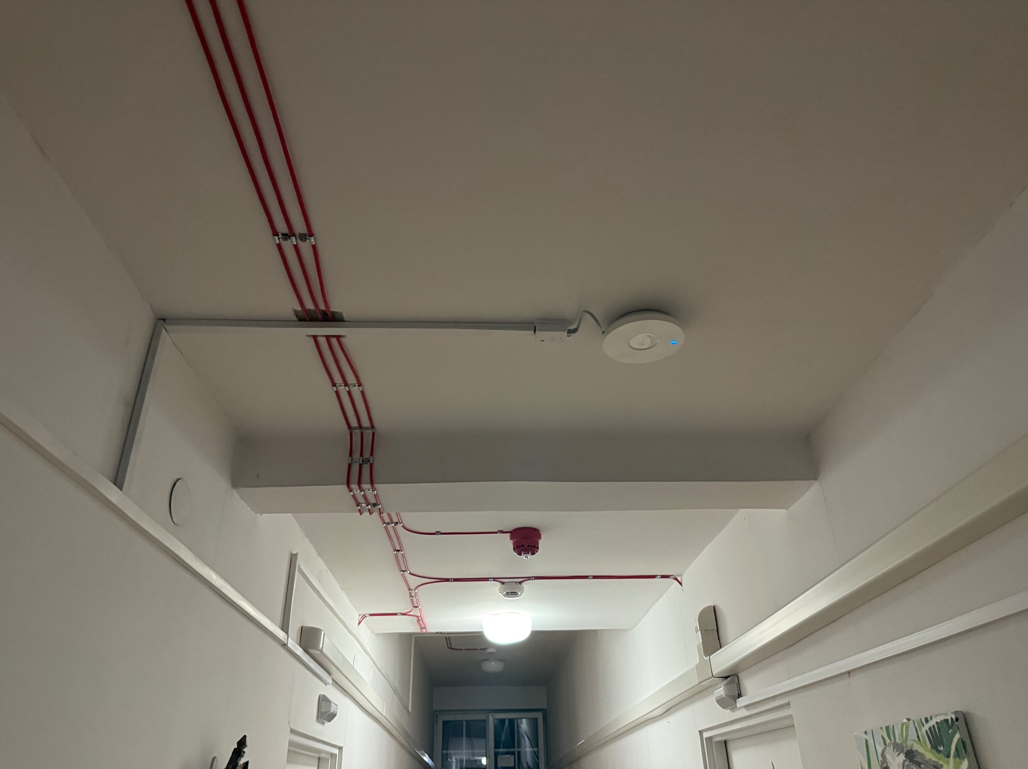 Korytarz z widocznymi czerwonymi kablami elektrycznymi przymocowanymi do sufitu za pomocą metalowych uchwytów, czujnikiem dymu i punktem dostępowym Wi-Fi.