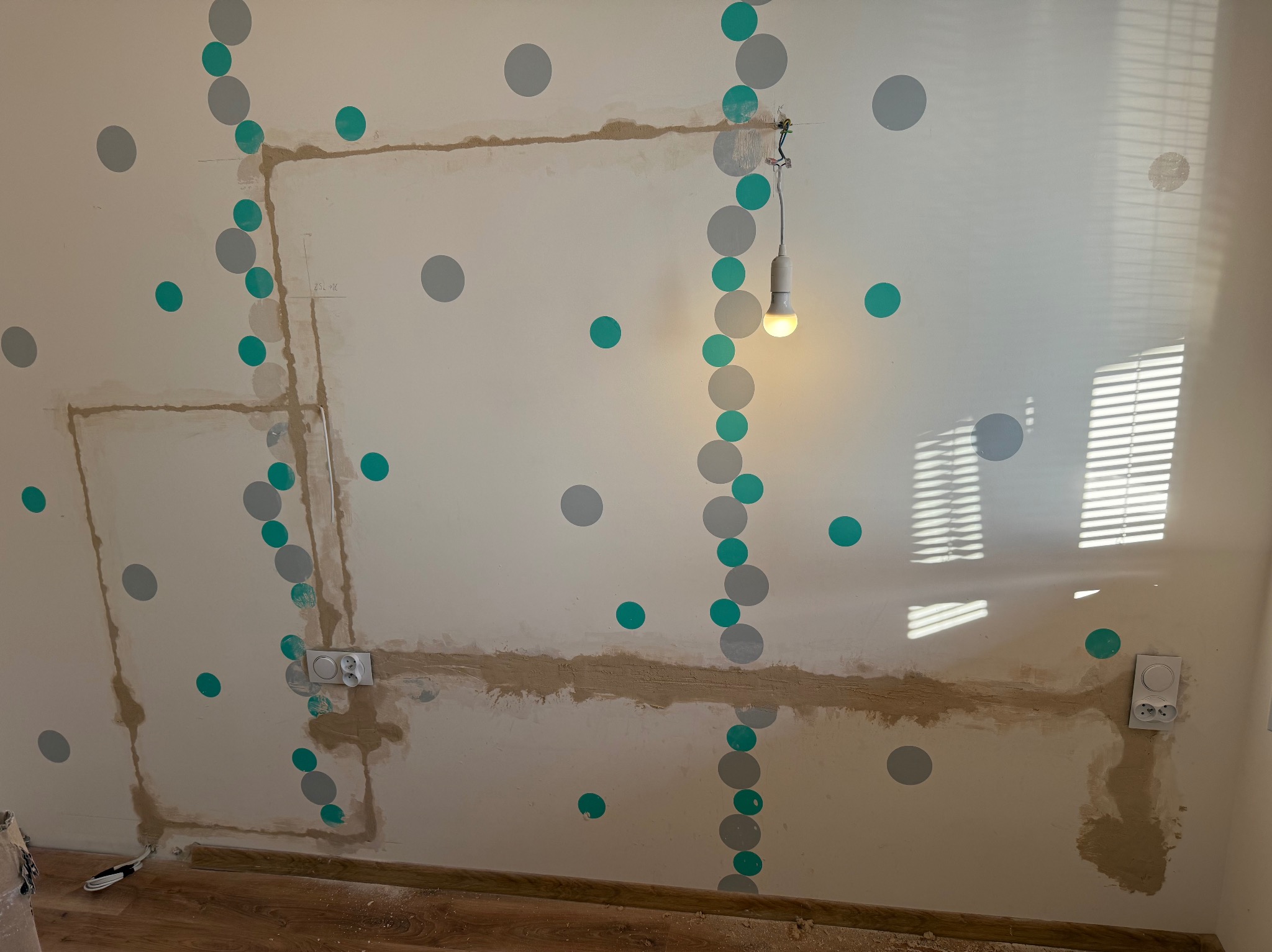 Ściana z wyciętymi bruzdami pod instalację elektryczną, widoczne gniazdka i przewody, na ścianie dekoracyjne kropki. Żarówka wisi na kablu.