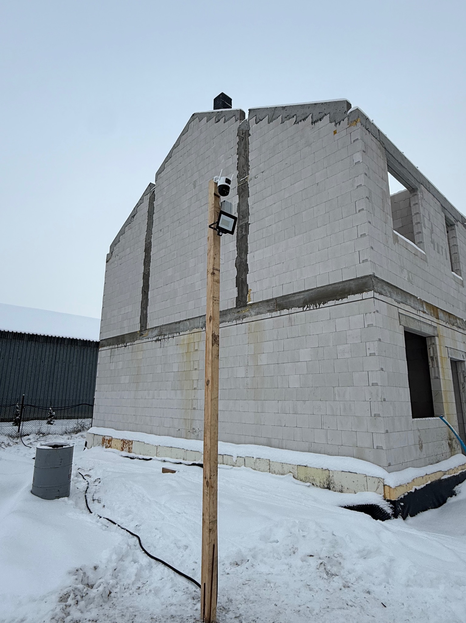 Niedokończony budynek z betonu komórkowego w zimowej scenerii. Widoczny tymczasowy słup z zamontowaną kamerą i reflektorem. Kabel biegnie po śniegu.