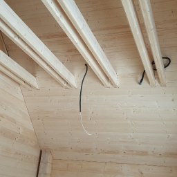 Atom - Wnętrze domu szkieletowego w trakcie budowy: drewniane ściany, strop z belkami i widoczne przewody instalacji elektrycznej. Surowy stan wykończenia, jasne drewno.