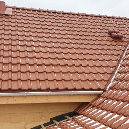 Atom - Czerwona dachówka ceramiczna na drewnianym domu szkieletowym, z widocznymi elementami systemu rynnowego i resztkami śniegu w załamaniach dachu.