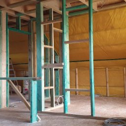 Atom - Wnętrze domu w trakcie budowy szkieletowej: widoczne drewniane ramy, ocieplenie ścian żółtą wełną mineralną, drabiny i narzędzia budowlane.