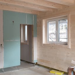 Atom - Wnętrze domu w trakcie wykańczania: drewniane ściany, zamontowane płyty karton-gips, widoczne instalacje elektryczne i narzędzia. Etap prac wykończeniowych.