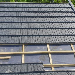 Atom - Montaż dachu z blachodachówki modułowej w kolorze grafitowym, widoczne okno dachowe i konstrukcja łatowania. Tło stanowią drzewa.