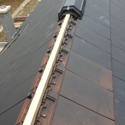 Atom - Montaż dachówki ceramicznej z drewnianą łatą wzdłuż kalenicy, widok z góry na fragment dachu w trakcie budowy, z kominem w tle i zielenią w oddali.