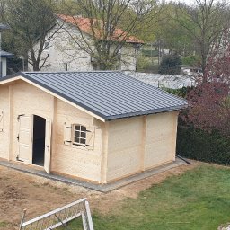 Atom - Drewniany domek letniskowy z szarą blachodachówką, otwartymi drzwiami i okiennicami, na trawniku z młodym drzewkiem. W tle dom z panelami słonecznymi.