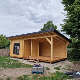 Atom - Drewniany domek letniskowy z czarną stolarką okienną i zadaszonym tarasem, na działce z zielenią i elementami budowlanymi. Widok z zewnątrz, pochmurne niebo.