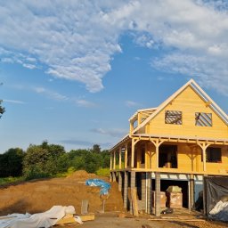 Atom - Wznoszenie domu szkieletowego na fundamencie z bloczków betonowych, widok z dołu na konstrukcję w surowym stanie, błękitne niebo w tle.