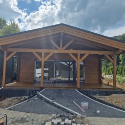 Atom - Drewniany domek letniskowy z tarasem w trakcie budowy, widoczne kostki brukowe i narzędzia. W tle zieleń i góry.