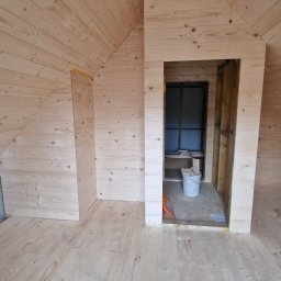 Atom - Poddasze wykończone jasnym drewnem, widoczne deski na podłodze, ścianach i suficie. W tle otwór drzwiowy z widocznym wiadrem i materiałami budowlanymi.