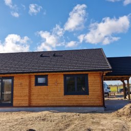 Atom - Drewniany dom z ciemną dachówką i czarnymi ramami okiennymi w słoneczny dzień. Widoczny fragment zadaszenia i teren budowy.