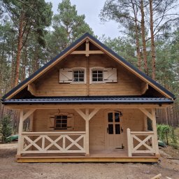 Atom - Drewniany domek letniskowy z gankiem, okiennice w stylu rustykalnym, czarny dach, otoczony lasem sosnowym. Widok z przodu, w pochmurny dzień.