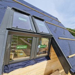 Atom - Montaż okien dachowych Fakro na drewnianej konstrukcji dachu, zabezpieczonej membraną Titanium Pro, widok z dołu pod kątem, w tle zieleń i błękit nieba.