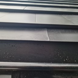 Atom - Czarny, metalowy panel elewacyjny z widocznymi łączeniami i kroplami wody, ukazujący detale wykończenia.