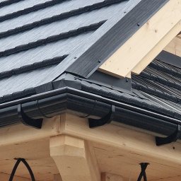 Atom - Fragment dachu z czarną blachodachówką i orynnowaniem, widoczne drewniane elementy konstrukcyjne. Detal wykończenia dachu domu szkieletowego.