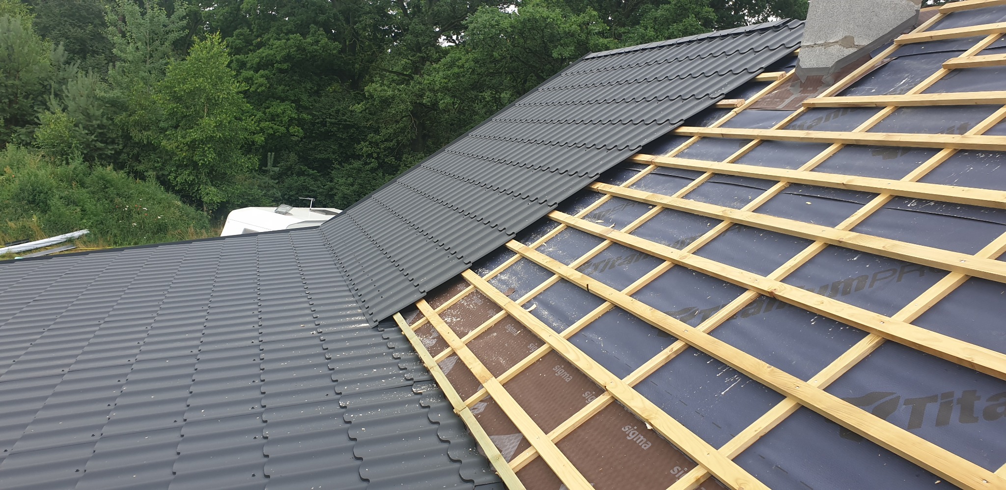 Częściowo pokryty szarą blachodachówką dach z widoczną konstrukcją drewnianą i membraną, komin w tle, zieleń drzew w tle.