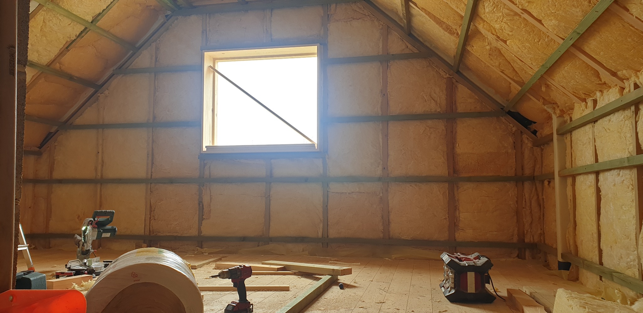 Poddasze z oknem w trakcie ocieplania wełną mineralną, widoczne drewniane elementy konstrukcyjne i narzędzia pracy.
