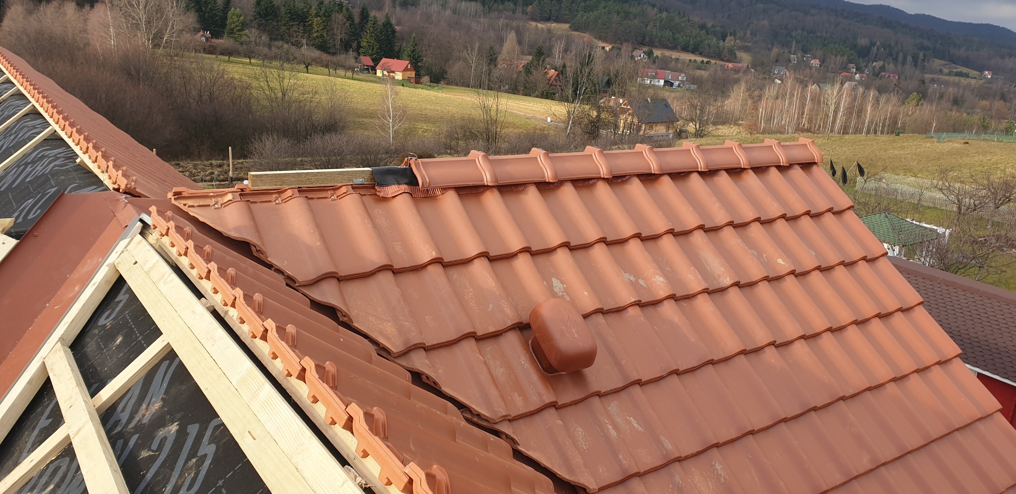 Częściowy widok na nowo kryty dach z brązowej dachówki ceramicznej, z widoczną konstrukcją drewnianą i membraną dachową. W tle pagórkowaty krajobraz.