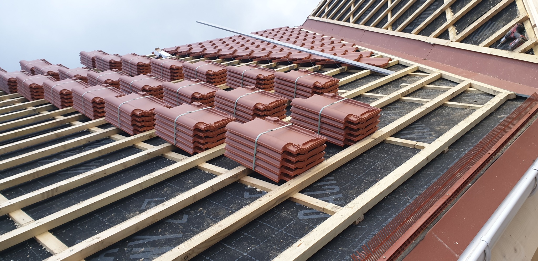 Układanie brązowej dachówki ceramicznej na drewnianej konstrukcji dachu pokrytej czarną membraną. Widoczne pakiety dachówek i elementy mocujące.