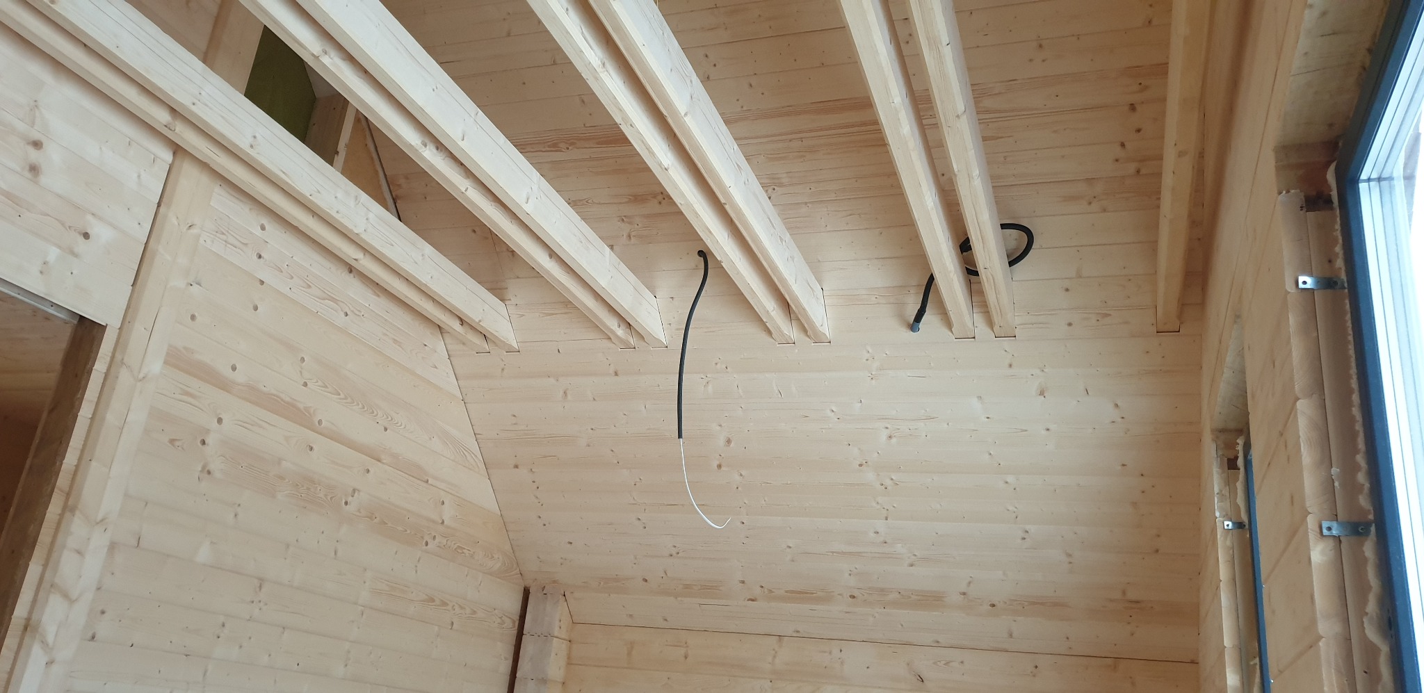 Wnętrze domu szkieletowego w trakcie budowy: drewniane ściany, strop z belkami i widoczne przewody instalacji elektrycznej. Surowy stan wykończenia, jasne drewno.