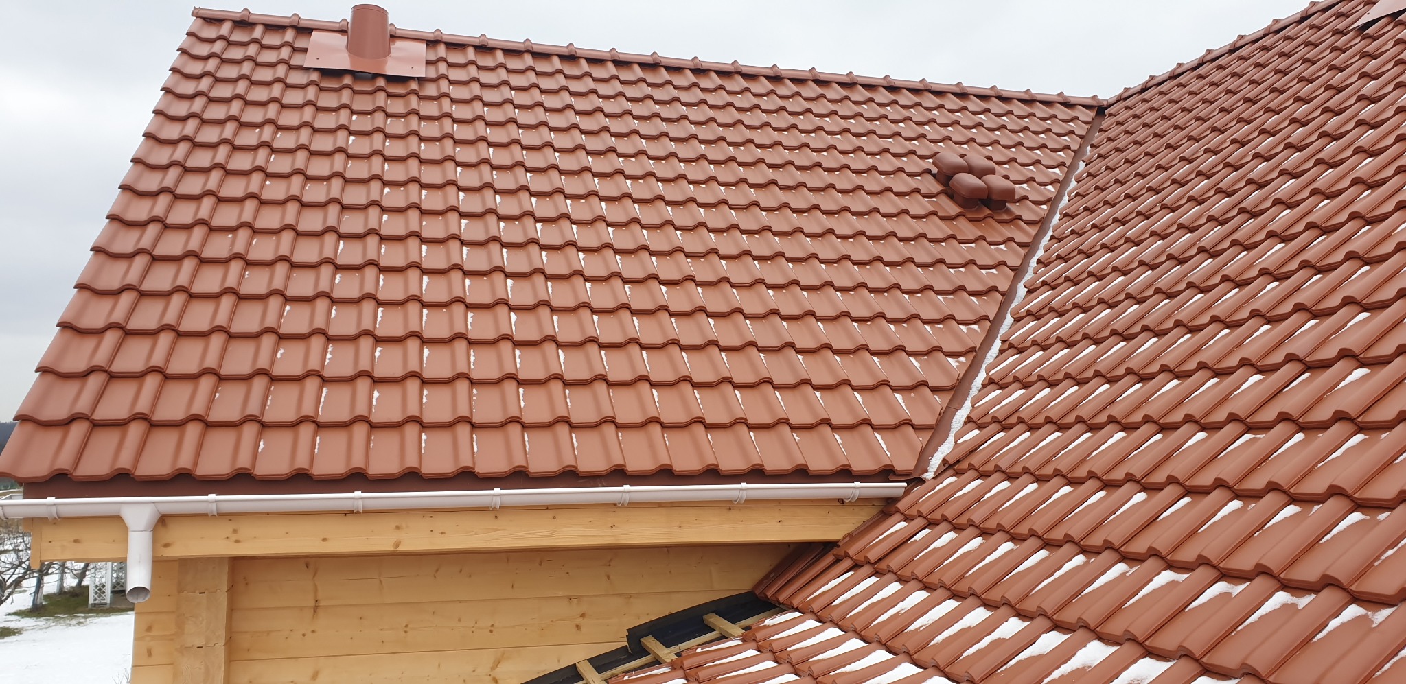 Czerwona dachówka ceramiczna na drewnianym domu szkieletowym, z widocznymi elementami systemu rynnowego i resztkami śniegu w załamaniach dachu.