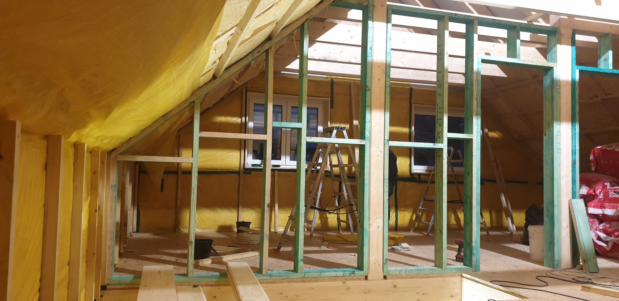Szkieletowa konstrukcja poddasza z oknami, ocieplona żółtą wełną mineralną, widoczne drabiny i narzędzia, etap prac wykończeniowych.