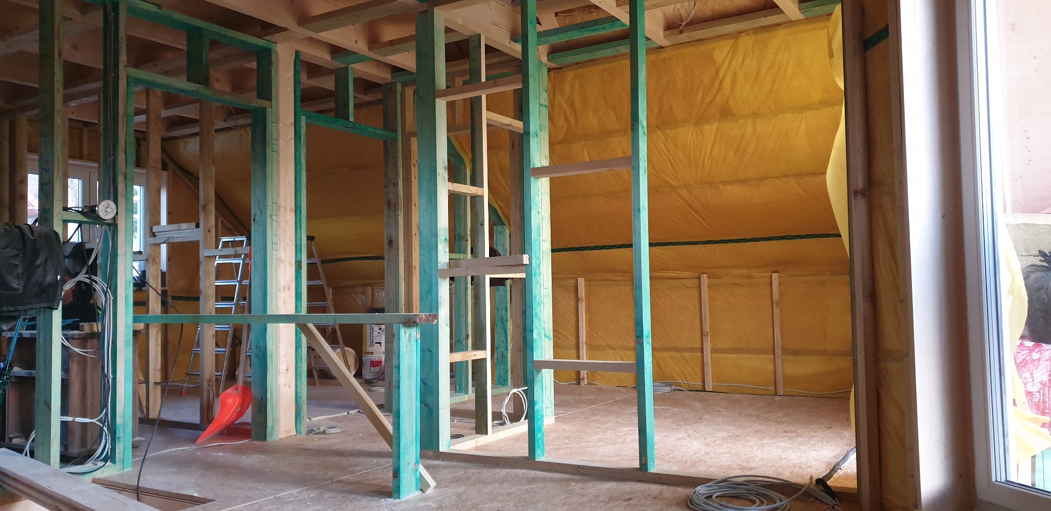 Wnętrze domu w trakcie budowy szkieletowej: widoczne drewniane ramy, ocieplenie ścian żółtą wełną mineralną, drabiny i narzędzia budowlane.