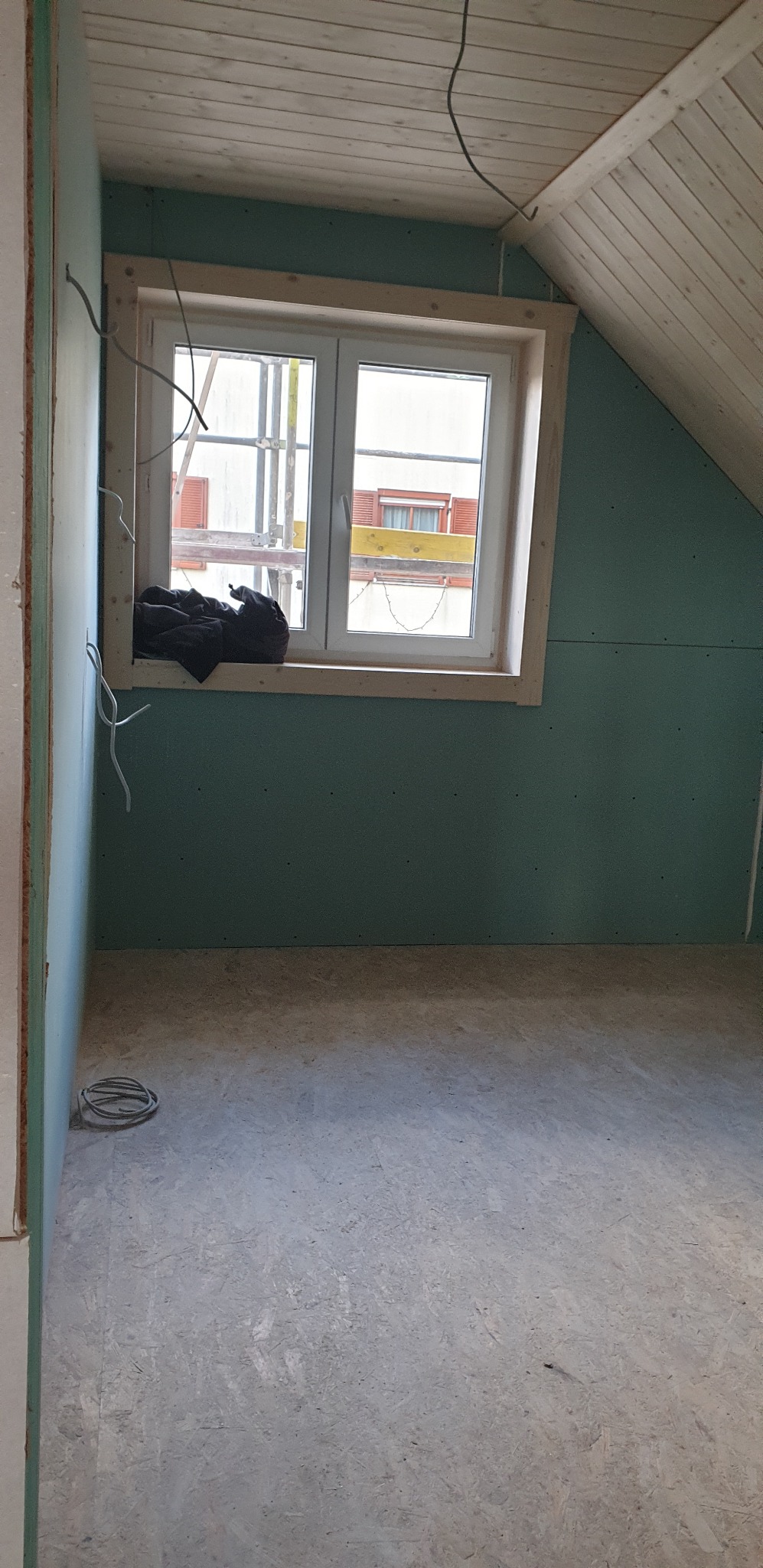 Poddasze w trakcie wykańczania: ściany z płyt karton-gips, drewniane okno z widokiem na rusztowanie, drewniana boazeria na skosie dachu, kable elektryczne.