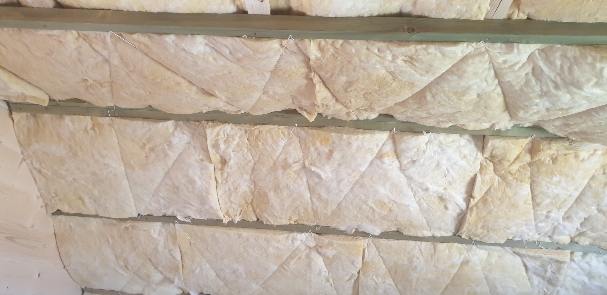Izolacja poddasza z wełny mineralnej, przymocowana sznurkiem do drewnianych belek stropowych. Widok z dołu na ocieplony strop.
