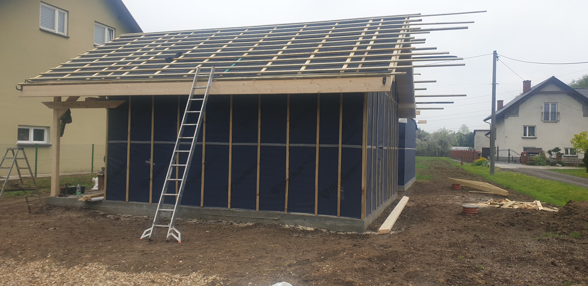 Budowa domu szkieletowego: widok na konstrukcję ścian z membraną i drewnianą więźbę dachową w trakcie montażu. Aluminiowa drabina oparta o ścianę.