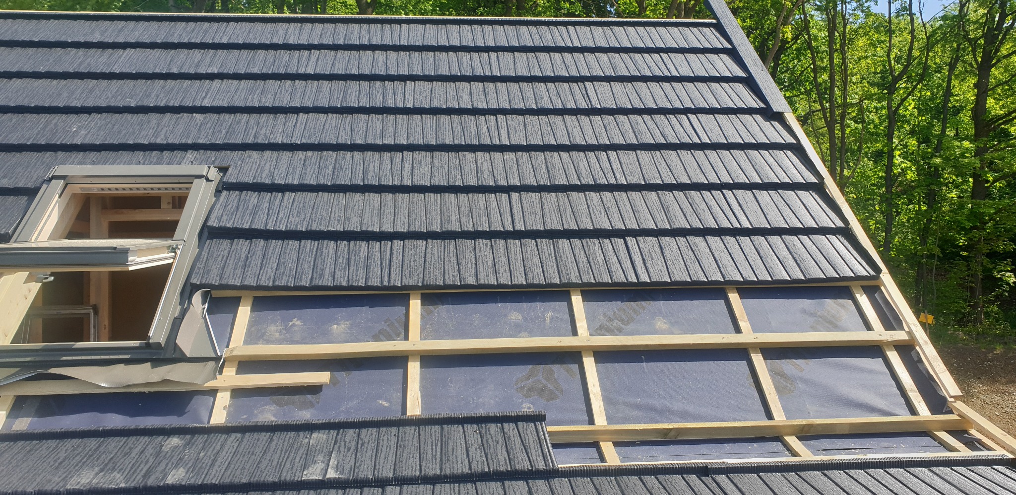 Montaż dachu z blachodachówki modułowej w kolorze grafitowym, widoczne okno dachowe i konstrukcja łatowania. Tło stanowią drzewa.
