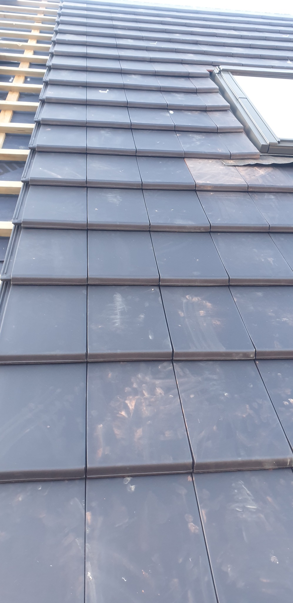 Ułożona dachówka ceramiczna w kolorze grafitowym z oknem dachowym. Widoczne drewniane elementy konstrukcji dachu. Ujęcie z dołu, podkreślające regularność wzoru.