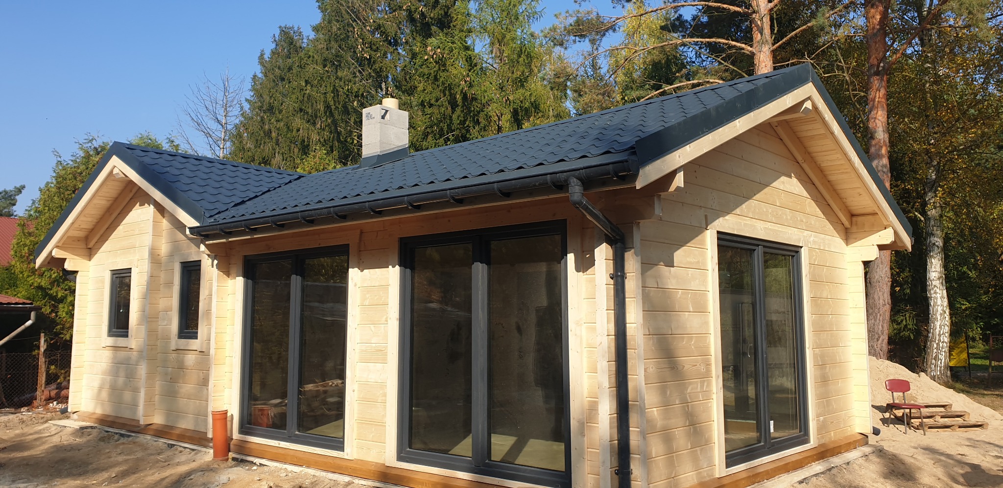 Nowoczesny dom szkieletowy z ciemną dachówką i dużymi oknami, otoczony zielenią. Widoczny fragment komina i rynny. Jasne drewno elewacji.