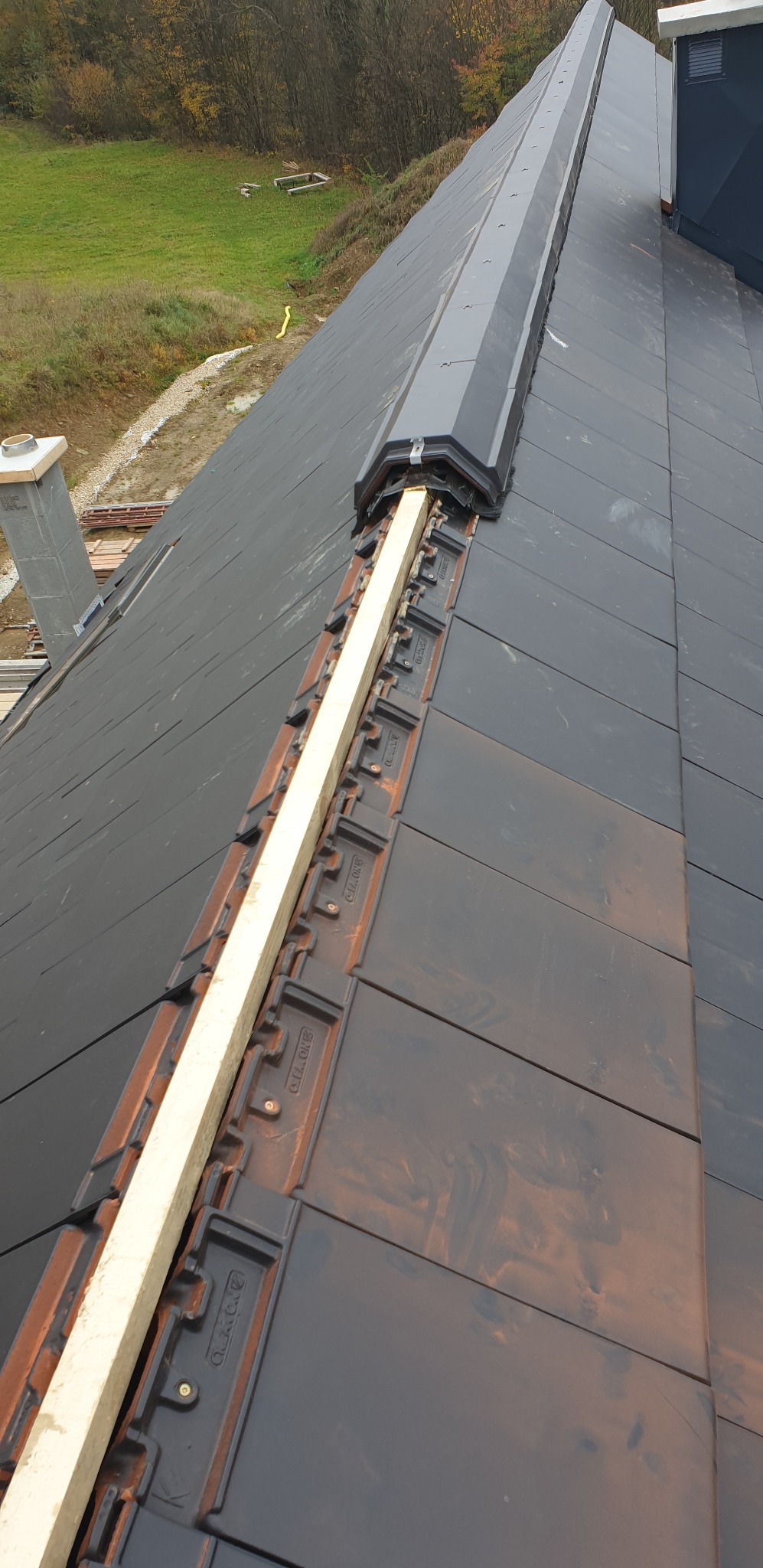 Montaż dachówki ceramicznej z drewnianą łatą wzdłuż kalenicy, widok z góry na fragment dachu w trakcie budowy, z kominem w tle i zielenią w oddali.