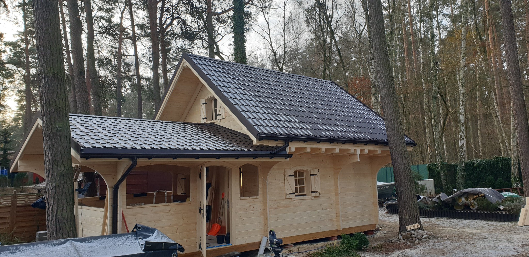 Drewniany domek letniskowy z ciemną dachówką w lesie, widok z zewnątrz, częściowo otwarte drzwi, narzędzia w środku, jasne drewno, czarne rynny.