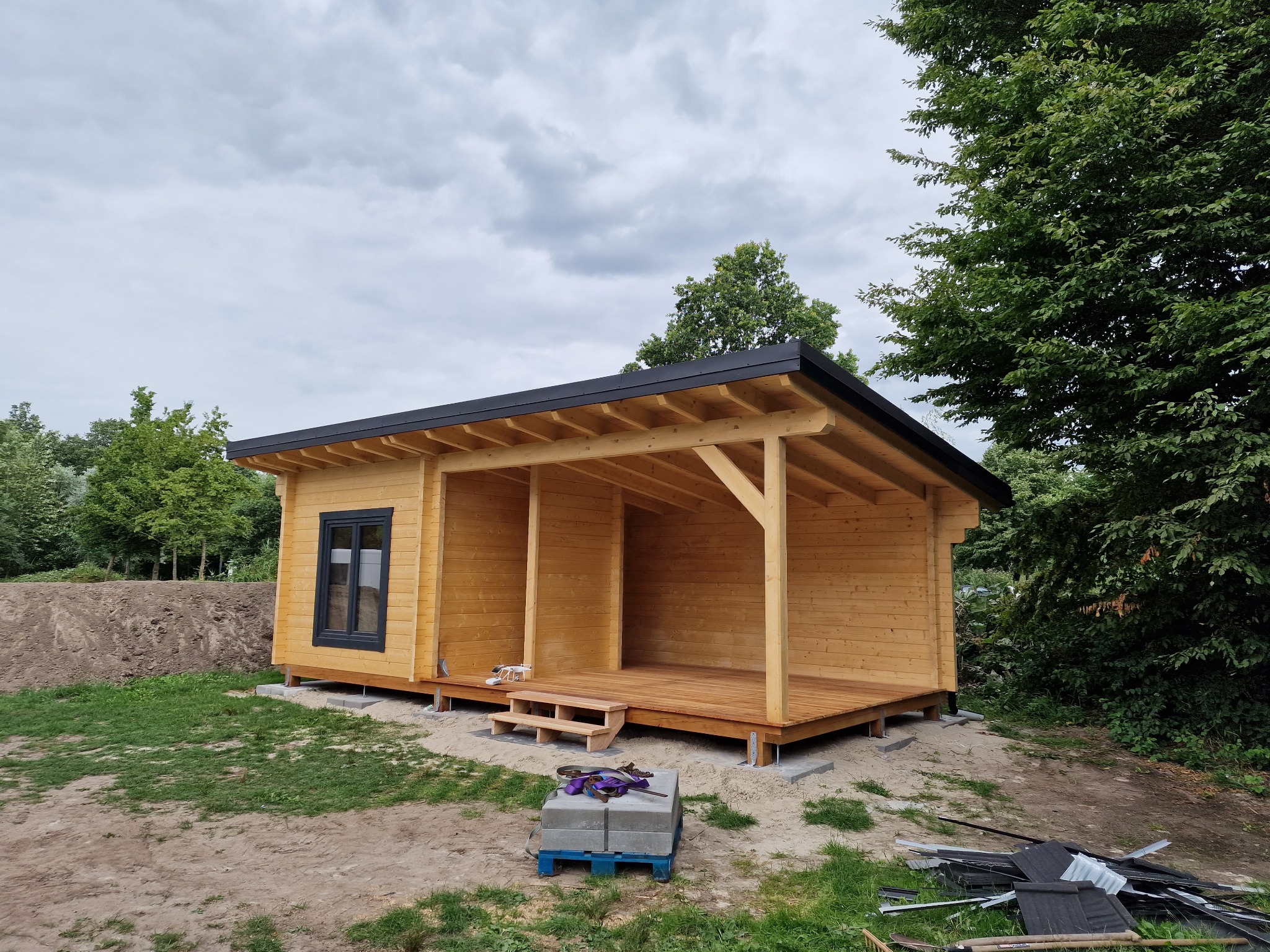 Drewniany domek letniskowy z czarną stolarką okienną i zadaszonym tarasem, na działce z zielenią i elementami budowlanymi. Widok z zewnątrz, pochmurne niebo.