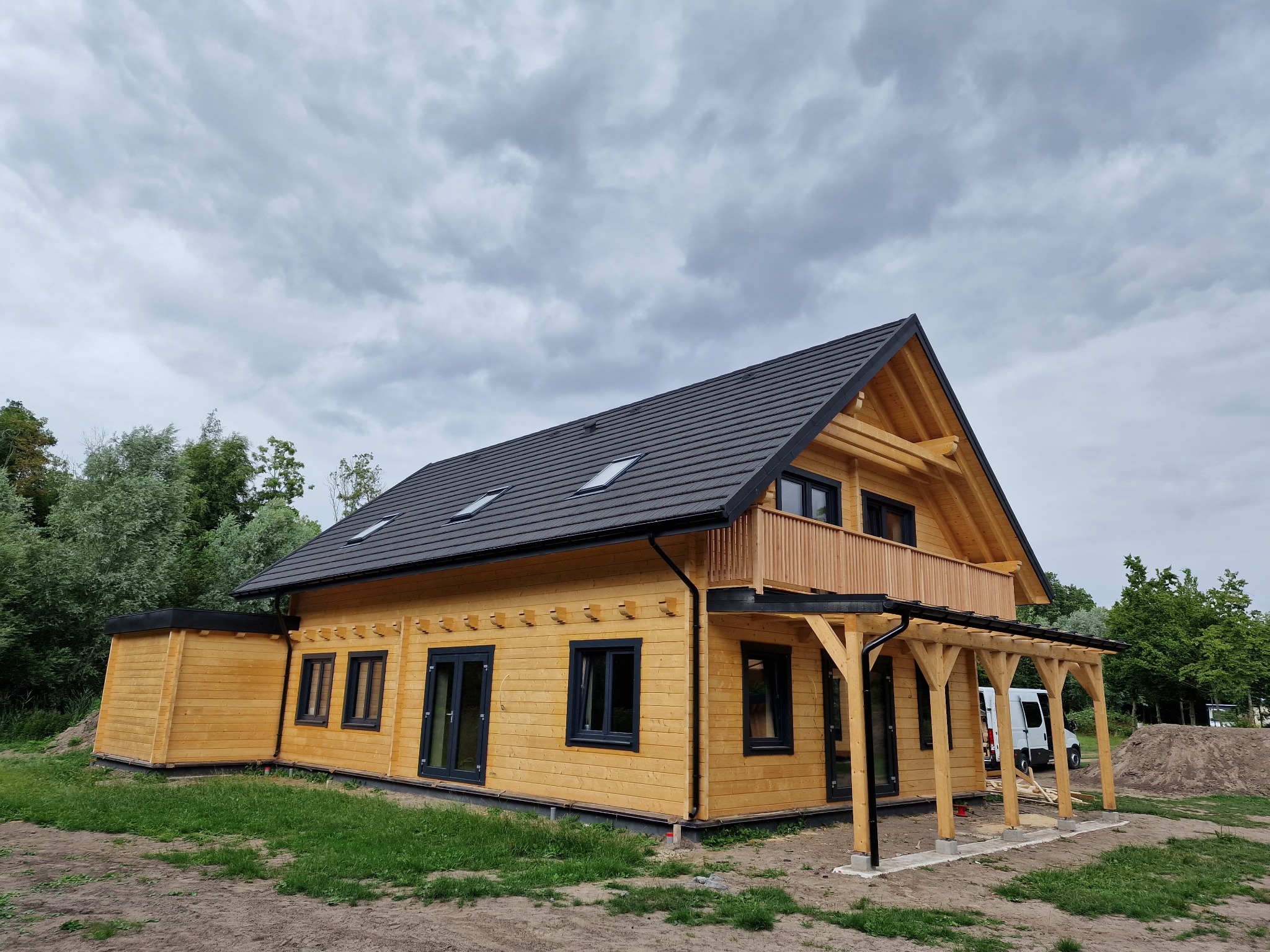 Nowoczesny dom szkieletowy z drewnianą elewacją, czarnymi oknami i dachem. Widoczna weranda i dobudówka. Pochmurne niebo w tle. Ujęcie z poziomu gruntu.
