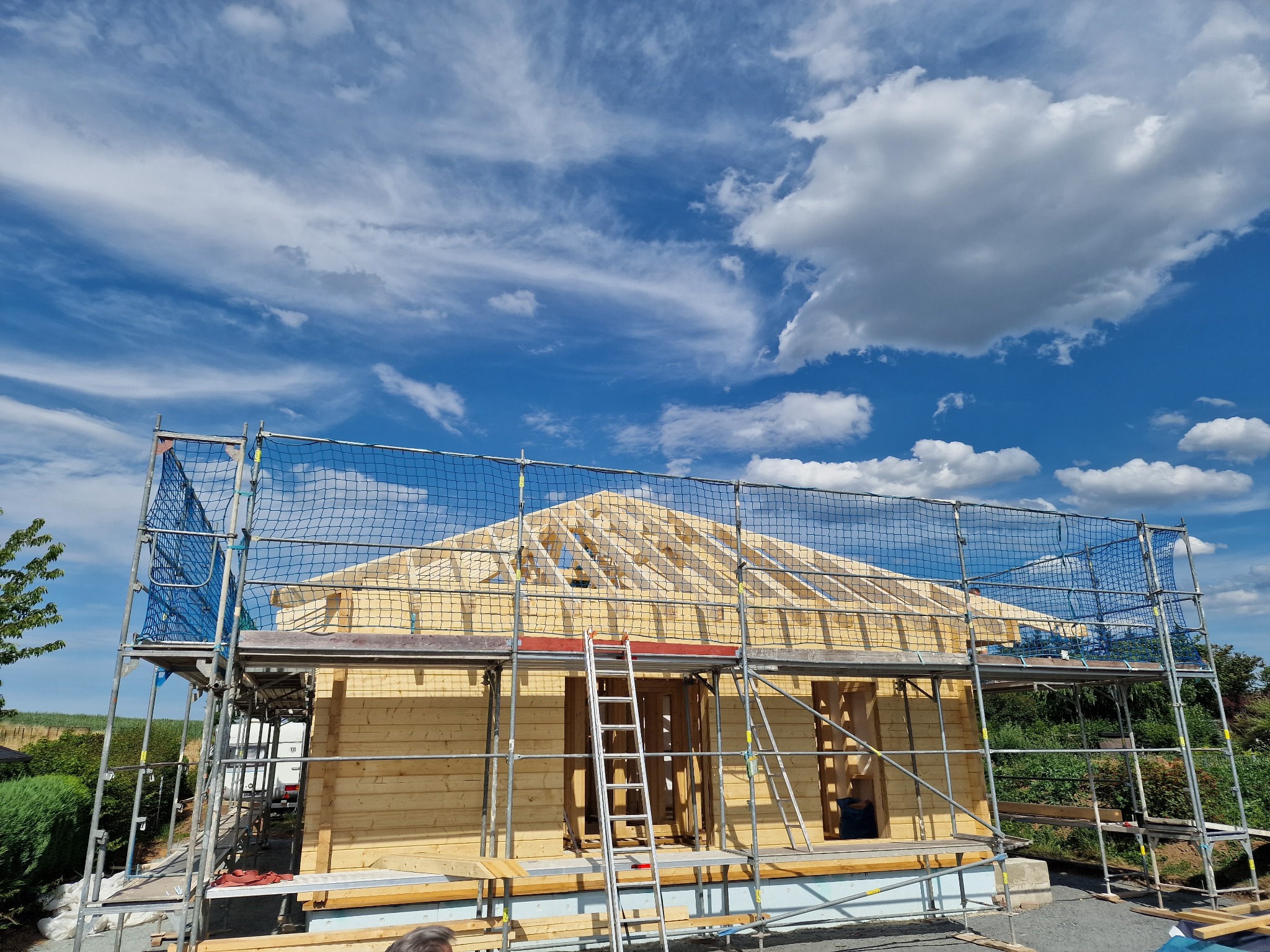 Wznoszenie drewnianego domu szkieletowego na fundamencie, widoczne rusztowania z siatką zabezpieczającą i konstrukcja dachu na tle błękitnego nieba z chmurami.