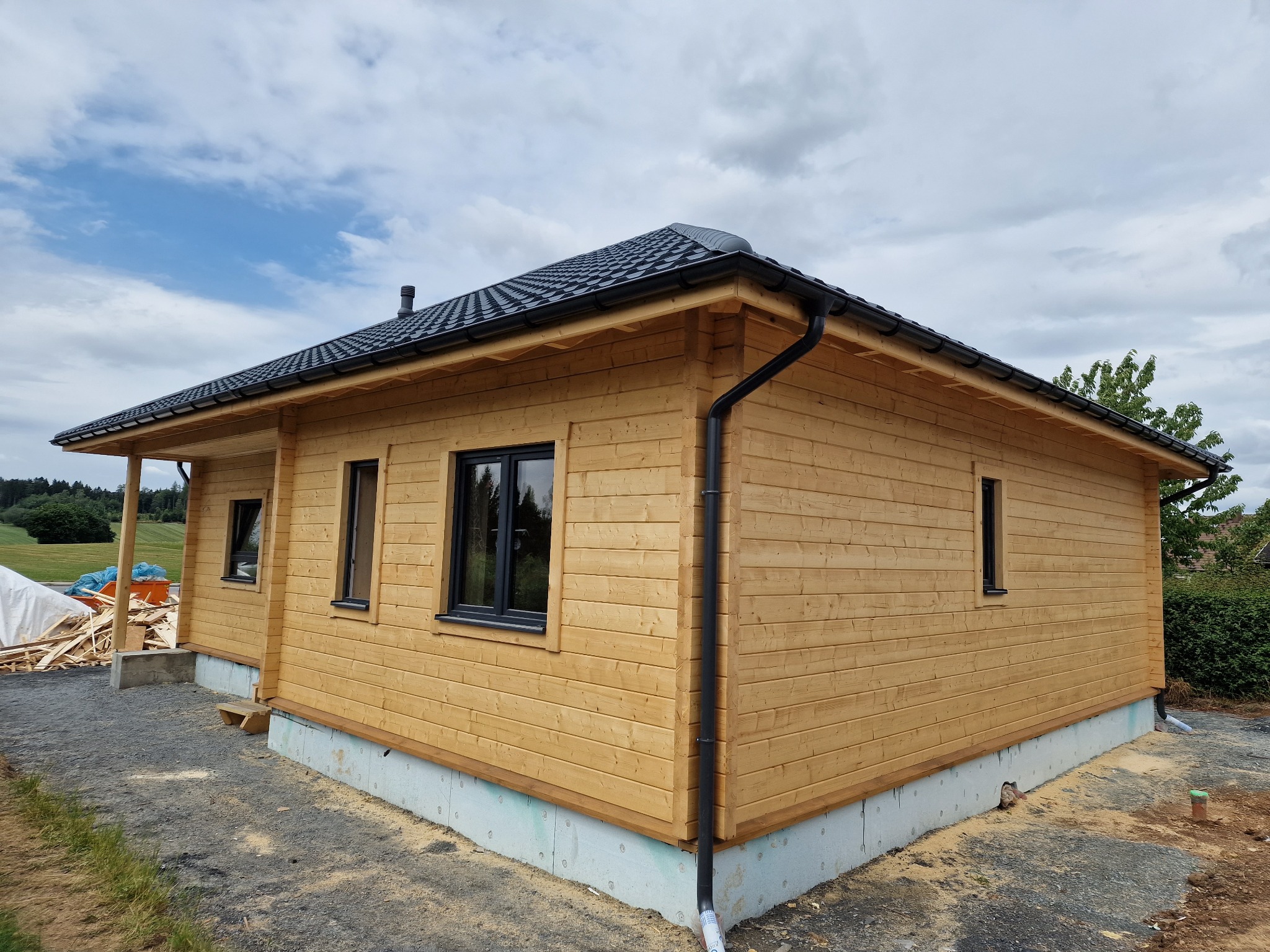 Niski, parterowy dom z drewnianych bali, z czarną dachówką i oknami. Widoczne fundamenty z izolacją. Częściowo wykończony, z elementami budowy w tle.